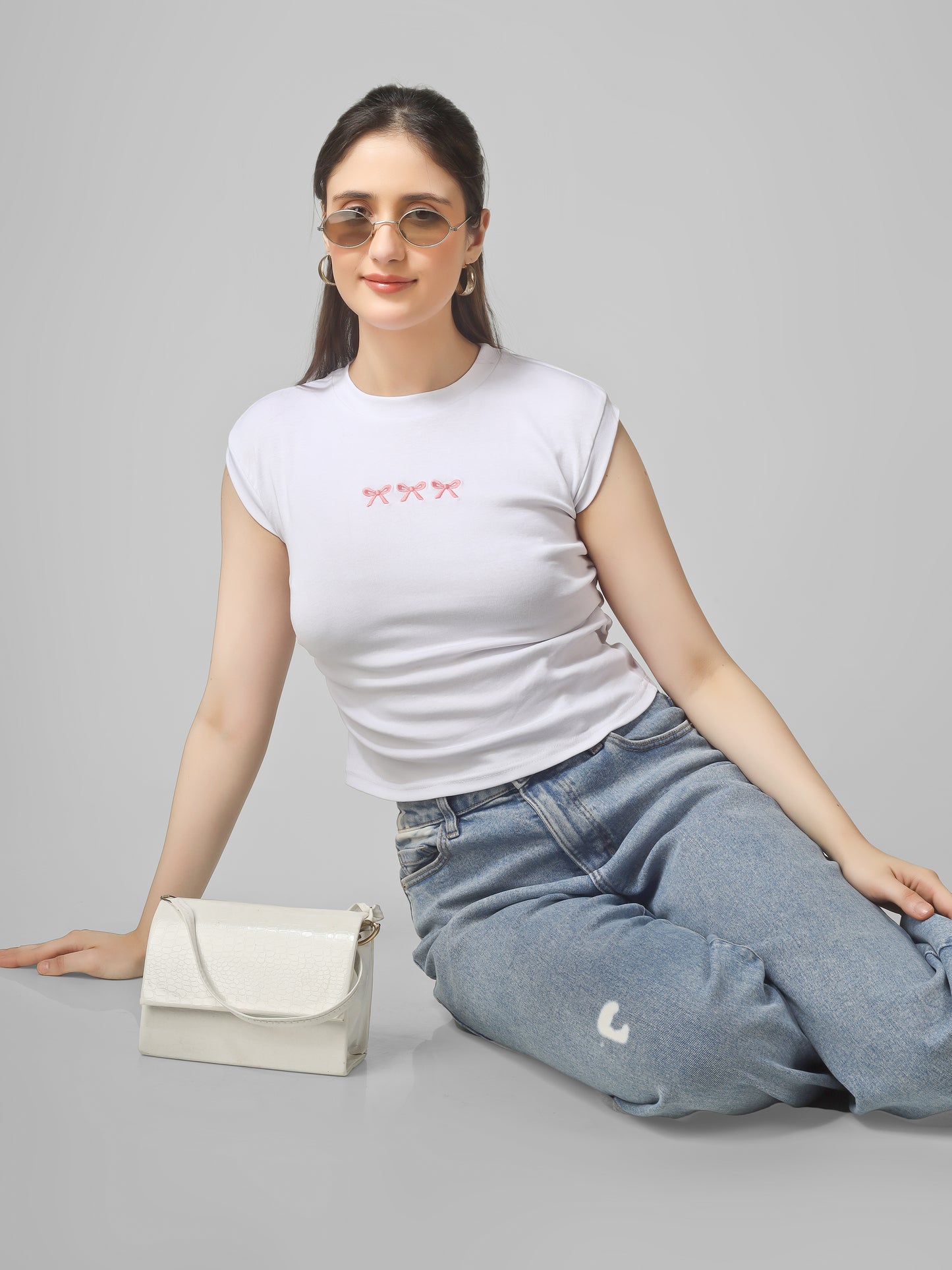 Woman wearing a white t-shirt with red text and blue jeans, sitting on the floor with a white handbag. Women white aesthetic crop top with bow embroidery slim fit